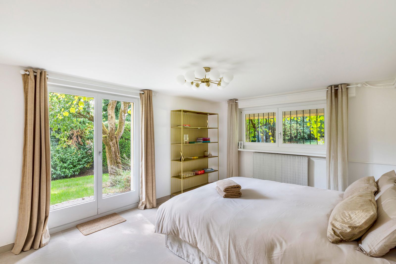 Chambre en rez-de-jardin, lit beige et étagère dorée face aux fenêtres